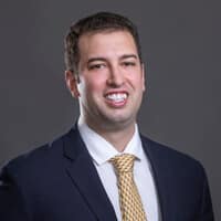 A person smiling in a professional portrait. They are wearing a navy suit, white shirt, and yellow tie against a dark gray background.