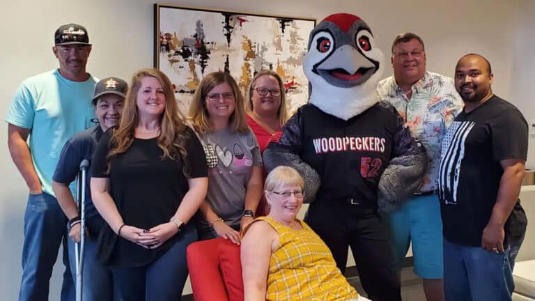 A group of eight people and a woodpecker mascot are posing happily indoors. One person is seated in a red chair.