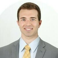A person smiling in a professional portrait. They are wearing a gray suit, light blue shirt, and yellow tie against a plain white background.