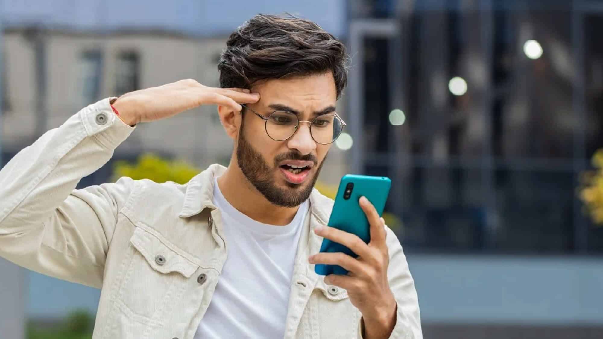 A man with glasses looks frustrated and confused while holding a smartphone, outdoors. He's pointing at his temple, suggesting disbelief.