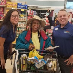 A smiling elderly woman in a colorful jacket and hat stands with a shopping cart filled with groceries. She's flanked by two credit union employees in blue uniforms inside a grocery store.