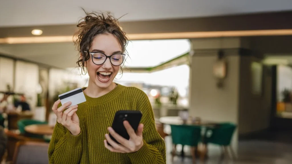 Happy woman looking at phone and holding credit card in other hand.