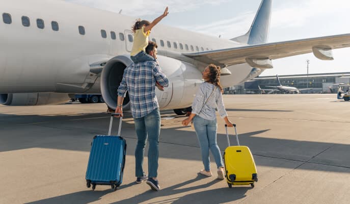 A family walks towards a plane on a sunny day. The father carries a child on his shoulders, both parents pulling colorful suitcases, conveying excitement.
