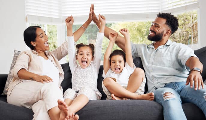 A joyful family of four sits on a couch, raising their arms in celebration. The parents flank two kids, all smiling widely in a bright, sunlit room.