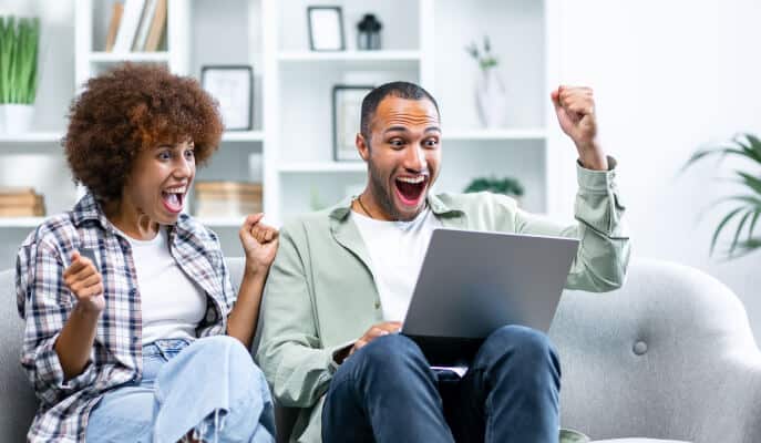 A joyful couple sits on a couch, excitedly celebrating in front of a laptop. Shelves with books and plants are visible in the bright room.