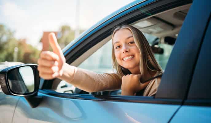 Smiling woman in a car gives a thumbs-up out of the window. Bright daylight reflects on the vehicle, indicating a positive, cheerful mood.