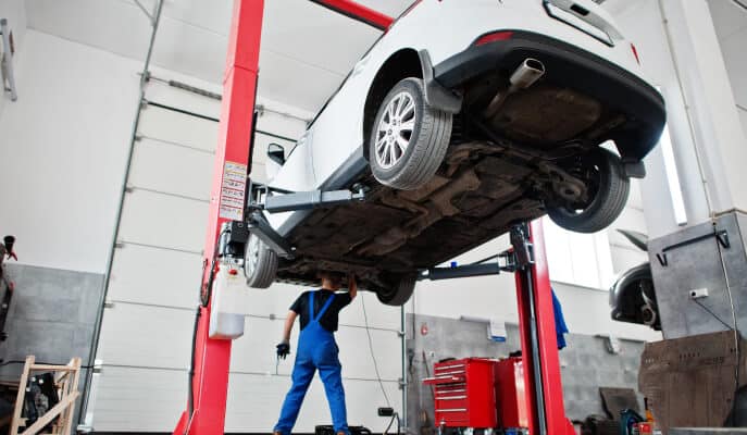A mechanic in blue overalls inspects the underside of a white car lifted on a red hydraulic lift in a spacious, bright garage, evoking a sense of professionalism.