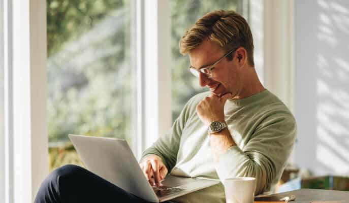 Man in glasses smiling at laptop; seated by sunlit window. Casual setting with a coffee cup, conveying a relaxed and focused atmosphere.
