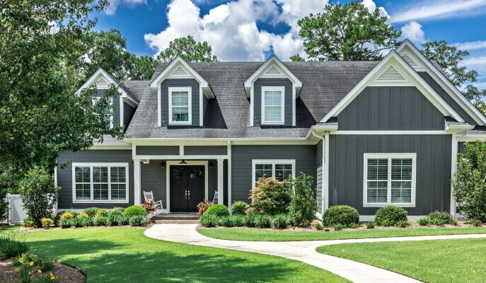 Grey two-story house with white trim, featuring a manicured lawn and a curved walkway. The house has dormer windows, a porch, and vibrant plants.