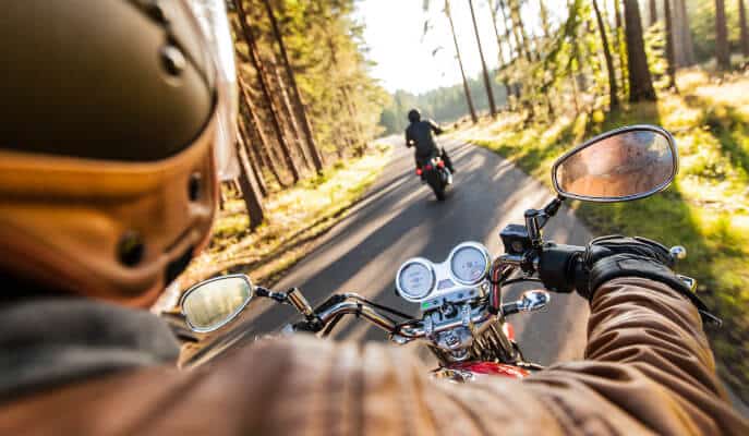 Two motorcyclists ride through a sunlit forest road. The foreground shows a rider's perspective, capturing handlebars and mirrored reflections.
