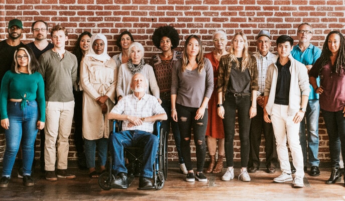 A diverse group of people stand together against a brick wall, including an elderly man in a wheelchair.