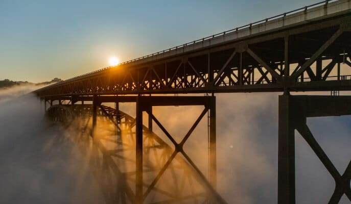 A metal bridge extends through thick fog, with the sun setting behind it, casting warm golden light and creating a serene, ethereal atmosphere.