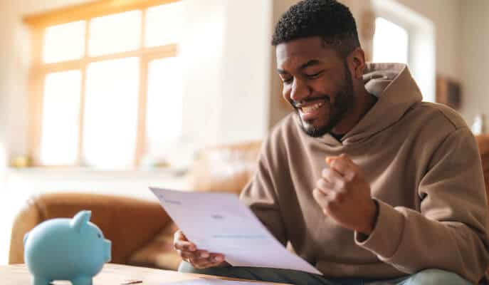 A man in a hoodie sits on a couch, smiling as he reads a document, with a blue piggy bank nearby. The scene conveys happiness and financial success.