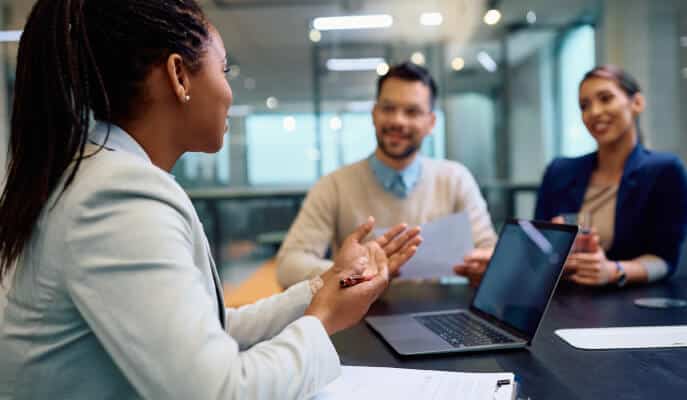A woman in a light blazer talks to a smiling man and woman across a table with a laptop, papers, and glass walls, creating a professional tone.