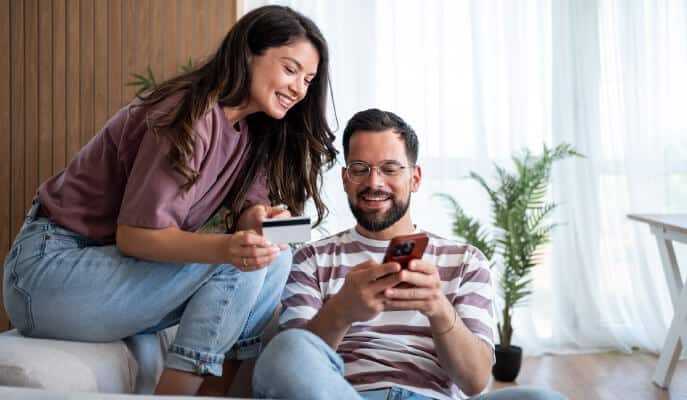 A smiling woman holds a credit card as she looks at a phone held by a bearded man. They're sitting in a bright, modern room with plants.