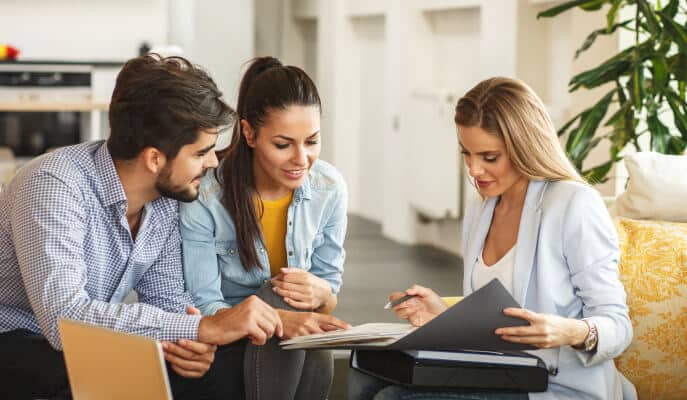 A young couple sits on a sofa, attentively listening to a woman in a blazer reviewing documents with them.