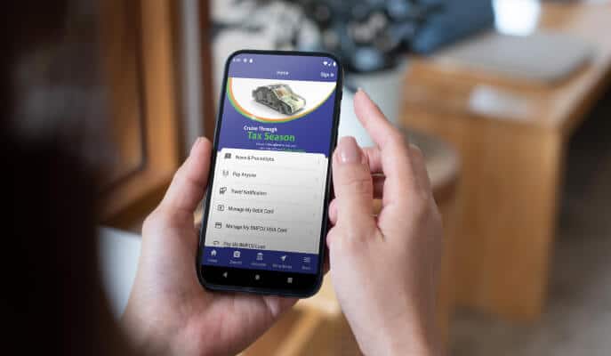 A person holds a smartphone displaying a financial app with options like “Manage My Debit Card.” The screen shows a tax season promotion. Cozy indoor setting.