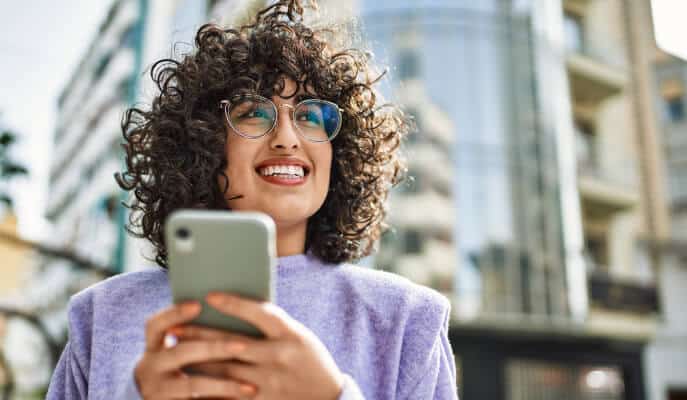 A smiling woman with curly hair and glasses holds a smartphone, wearing a purple sweater. She stands outdoors, with modern buildings in the background.