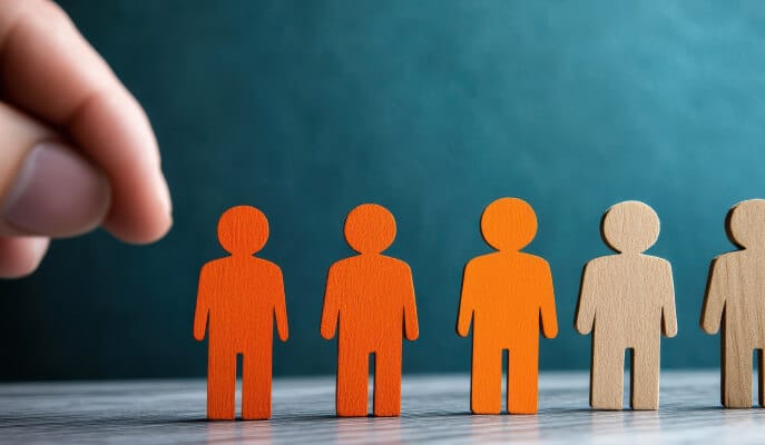 A hand is about to pick one of four orange wooden figures among two beige ones on a wooden surface. The background is teal, conveying choice and selection.