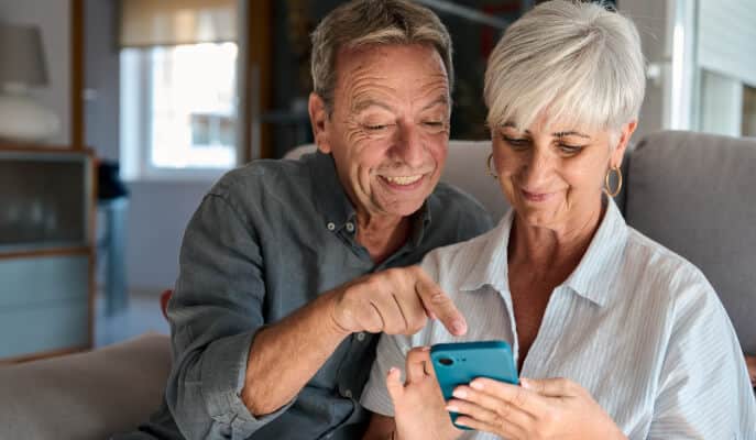 Elderly couple smiling warmly as they view a smartphone together on a sofa.