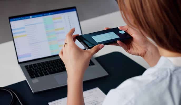 A person photographs a document with a smartphone at a desk. A laptop displaying a colorful spreadsheet is open in the background, suggesting a work setting.