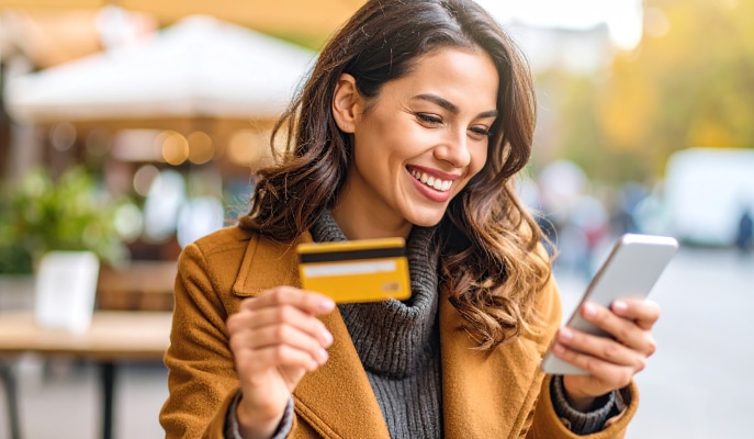 A smiling woman in a brown coat holds a credit card and smartphone outdoors. She appears happy and engaged, with soft-focus background lighting.
