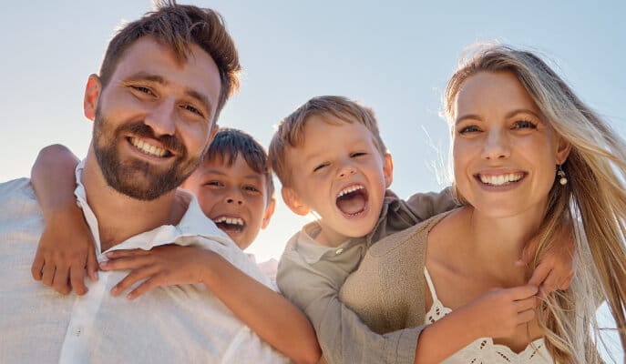 A joyful family of four poses outdoors, smiling widely under a clear blue sky. The parents hold their laughing children on their backs, conveying happiness.