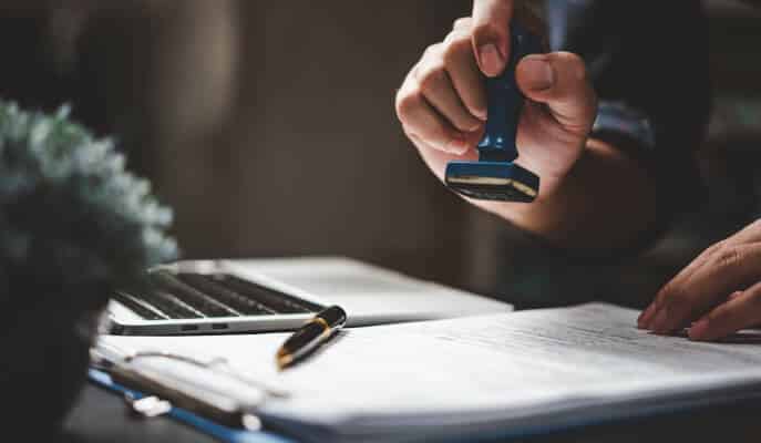 A person stamps a document beside a laptop, emphasizing official approval. A pen and plant are nearby, creating a focused, professional ambiance.