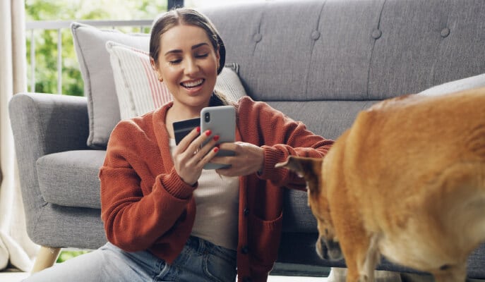 A woman in an orange sweater sits on the floor by a sofa, smiling at her phone while holding a credit card. A dog stands nearby, creating a cozy, joyful atmosphere.