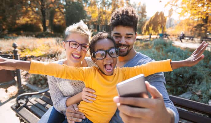 A joyful family takes a selfie on a park bench during autumn. The child, in a yellow sweater, spreads arms wide, excitedly smiling.