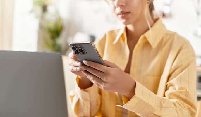 Woman in yellow button down shirt working at desk in front of laptop while looking at cell phone screen in hands.