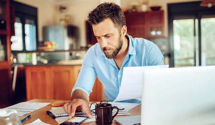 Man sitting at kitchen table looking intently at phone screen while surrounded with laptop, coffee mug and scattered documents.