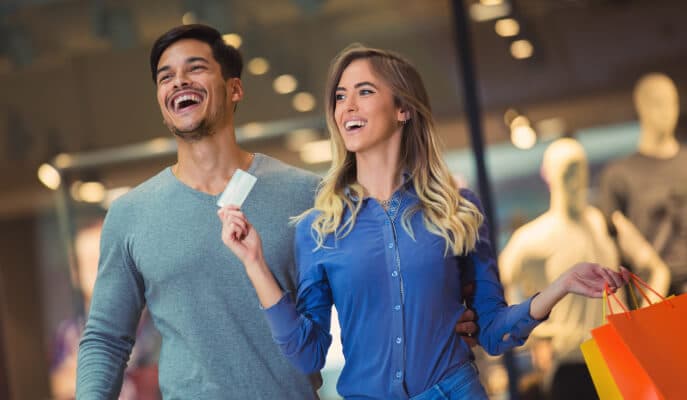 A smiling couple shopping in a mall, with the woman holding colorful bags and a credit card.