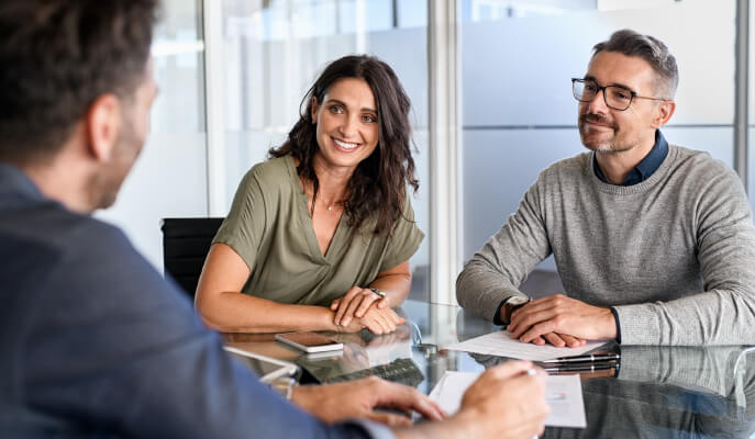 A woman and a man sit together at a glass table, smiling and attentively listening to a person across from them. Papers, a pen, and a smartphone rest on the table, suggesting a professional meeting or consultation in a bright, modern office.