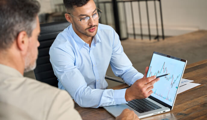 One man explaining to another charts on screen of laptop while they both sit at a large table.