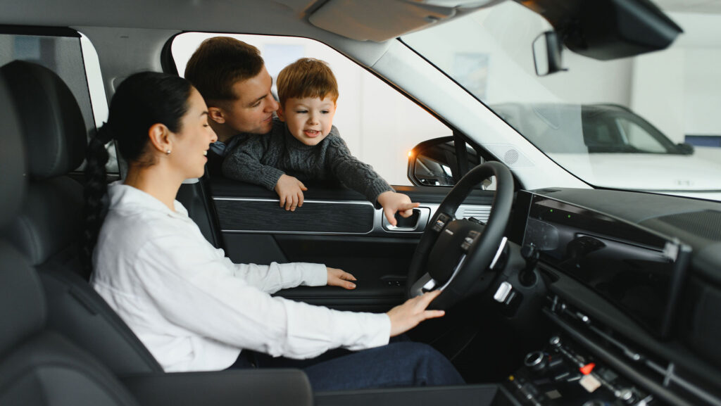 Woman test driving car while husband and child lean in front the driver's seat window.