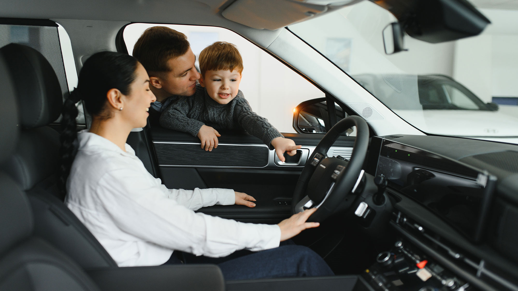 Woman test driving car while husband and child lean in front the driver's seat window.