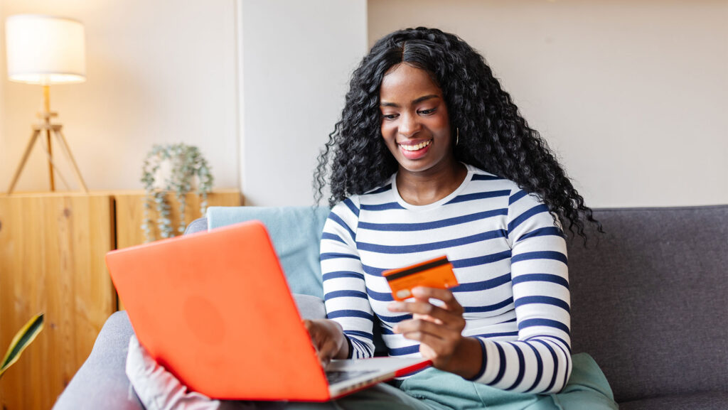 Young woman with black curly hard and blue and white striped shirt holding orange laptop and credit cards in front of her while sitting on couch in living room.