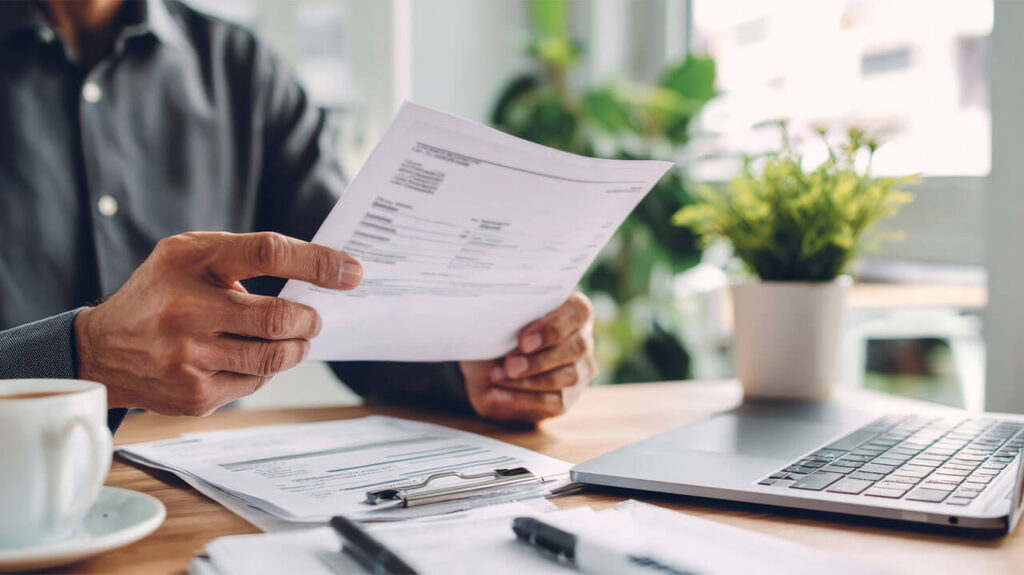 Man holding documents in hand with others laying in front of him on desk along with laptop, cup of coffee and plant.