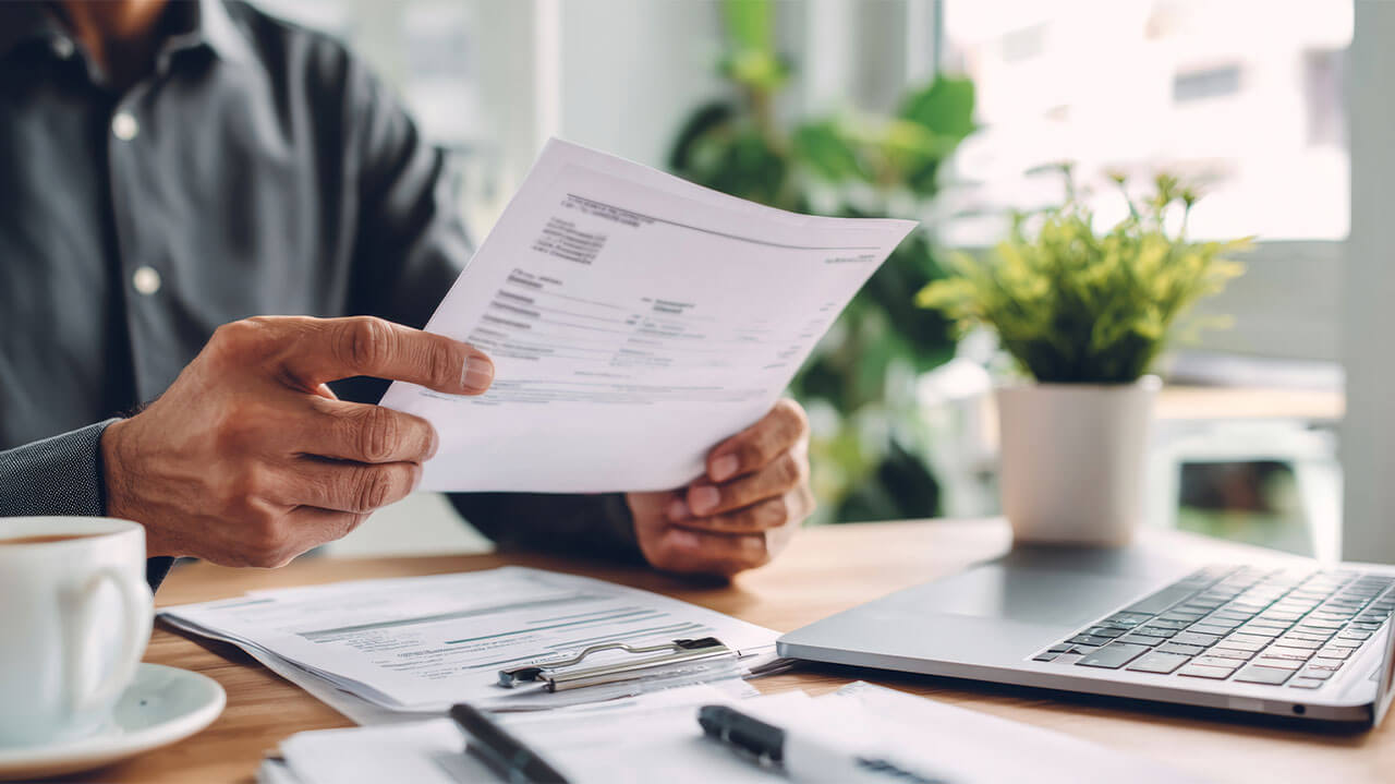 Man holding documents in hand with others laying in front of him on desk along with laptop, cup of coffee and plant.