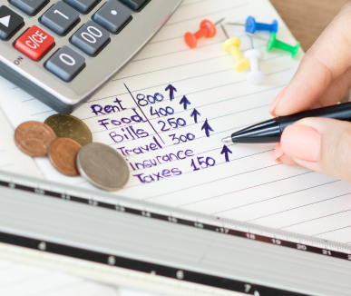 A hand writes on a lined notebook, listing expenses like rent and food with upward arrows. Nearby are coins, pushpins, and a calculator, conveying budgeting.