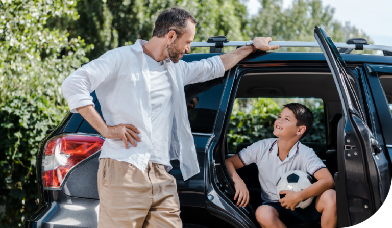 A man in a white shirt leans on an SUV, smiling at a boy with a soccer ball sitting inside. The scene is outdoors, suggesting a warm, cheerful atmosphere.
